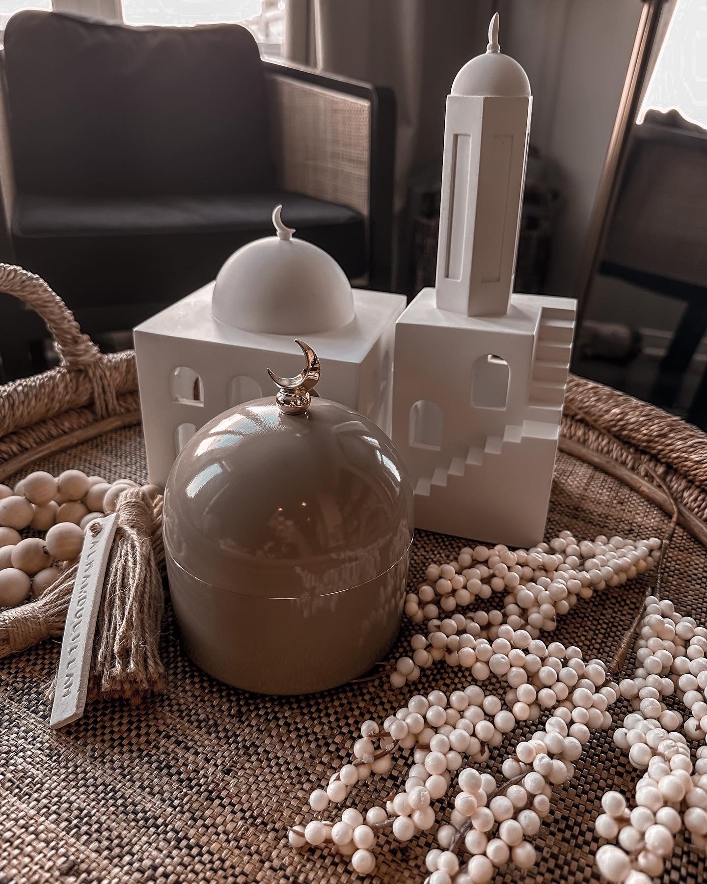 Dome shaped jar with crescent moon handle/topper, Ceramic mosque & minaret decorative statues, and some large, wooden tasbih beads, inside a shallow, round, woven basket atop a coffee table.