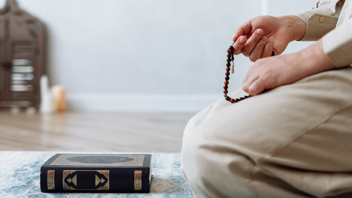 Man sitting on a carpet with a closed Quran in front of him and holding tasbih beads.